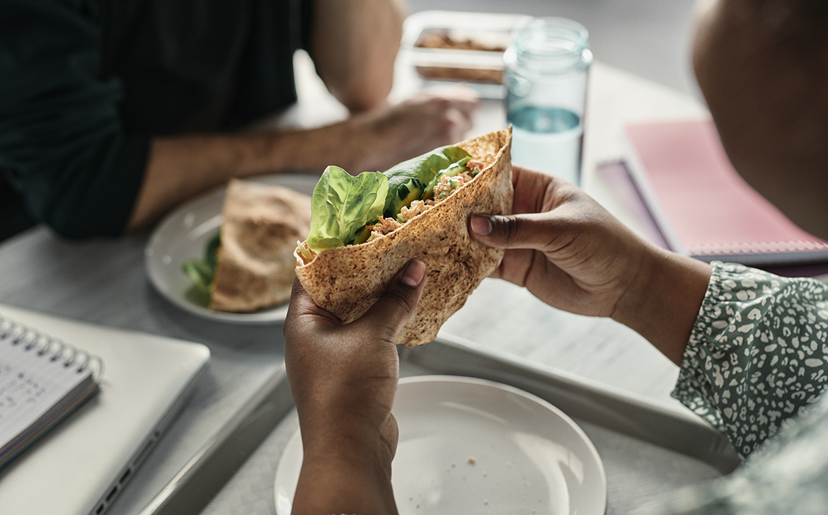 Une personne est assise dans une cafétéria et tient un sandwich pita fait de grains entiers. Un verre d’eau est posé à côté du repas comme boisson. Une autre personne est assise en face d’elle, avec un sandwich pita de grains entiers dans une assiette.